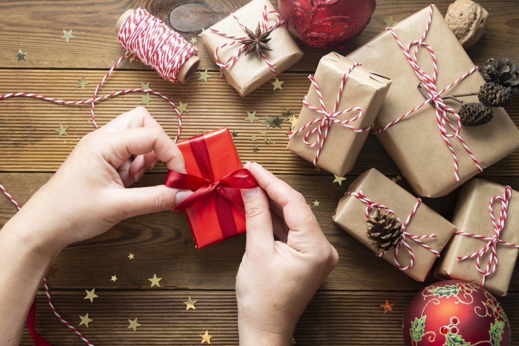 Female hands holding, wrapping Christmas gift box. Group of gift boxes wrapped in craft paper, red baubles, glitter over wooden tables. Christmas flat lay.